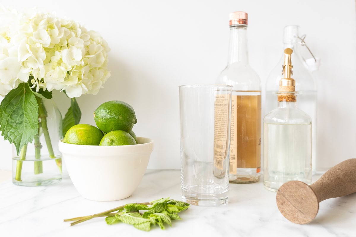 A kitchen counter with limes in a bowl, fresh mint, hydranges, and various cocktail-making supplies including bottles and a mixing glass for preparing vodka mojitos.