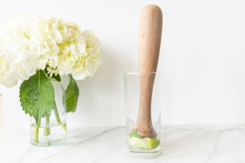 A wooden muddler in a glass with lime wedges beside a vase containing white hydrangeas on a marble countertop, ready to prepare a vodka mojito.