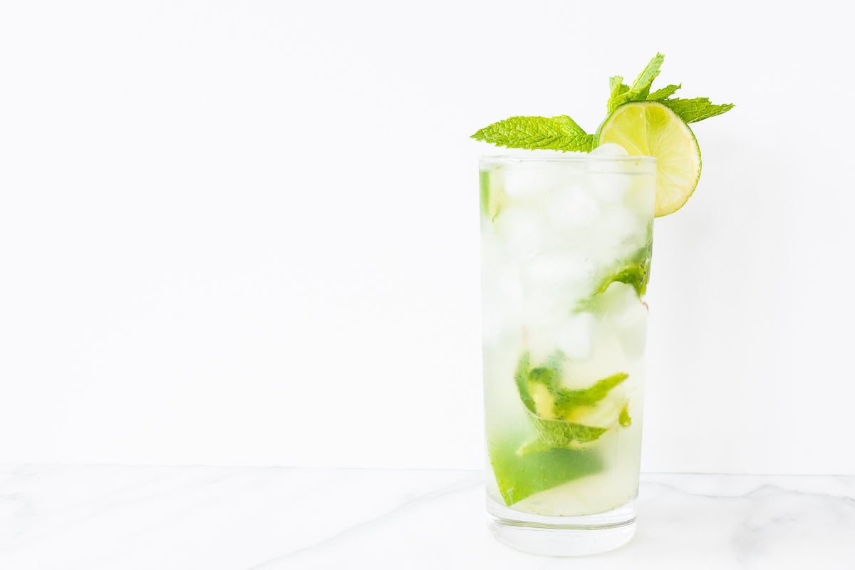 A glass of vodka mojito with lime slices and mint leaves against a white backdrop.