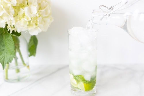 Water being poured from a clear bottle into a glass filled with ice cubes, lime slices, and mint leaves to make a Vodka Mojito, with a hydrangea bouquet in the background.