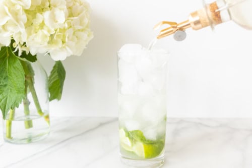 Pouring a clear vodka mojito into a glass with ice and lime slices, next to a vase of white hydrangeas on a marble countertop.