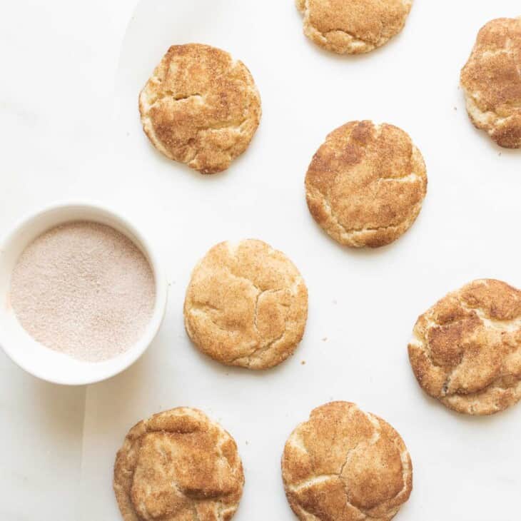 Soft and fluffy snickerdoodle cookies on a white countertop with a bowl of cinnamon and sugar. #easysnickerdoodlecookierecipe #softsnickerdoodlerecipe