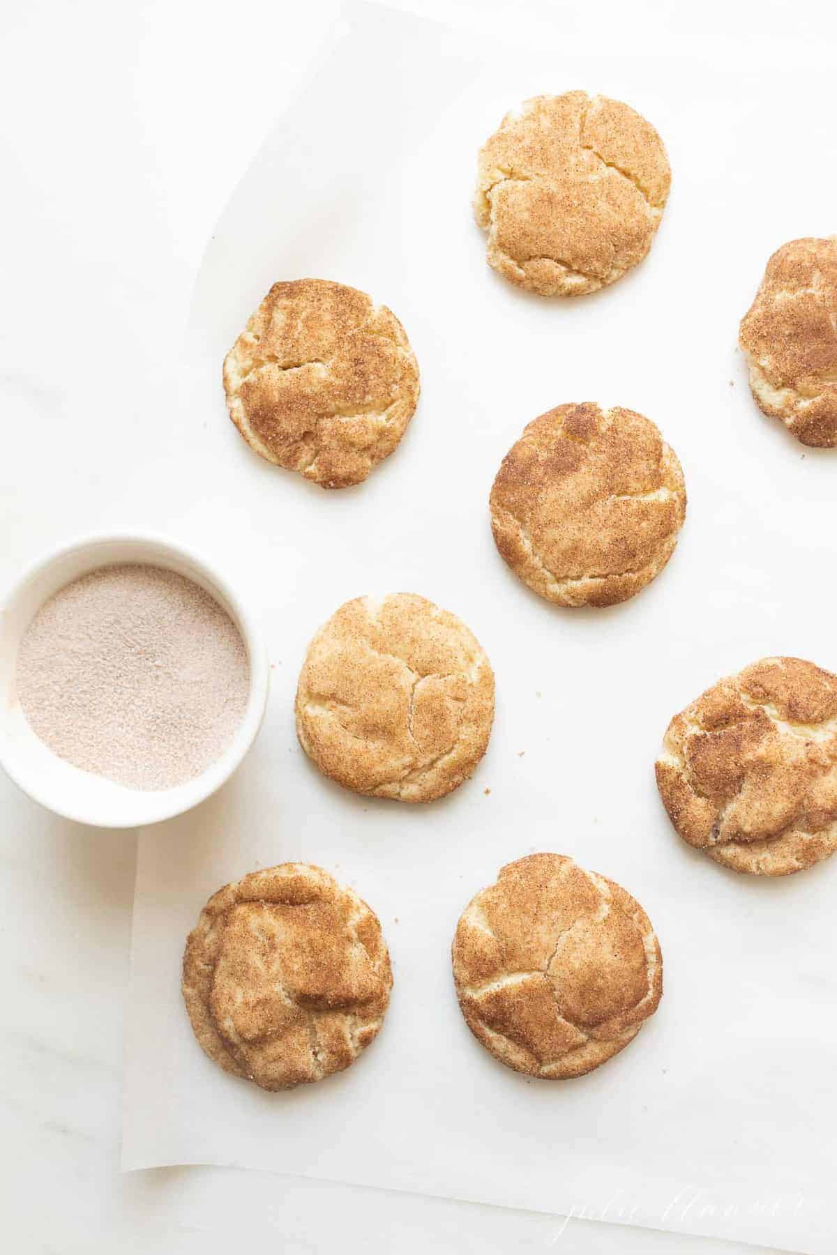 Snickerdoodle cookies on a white surface with a bowl of cinnamon sugar to the side. 