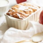 A small white ceramic loaf pan holding fresh baked apple cinnamon bread, drizzled with icing.