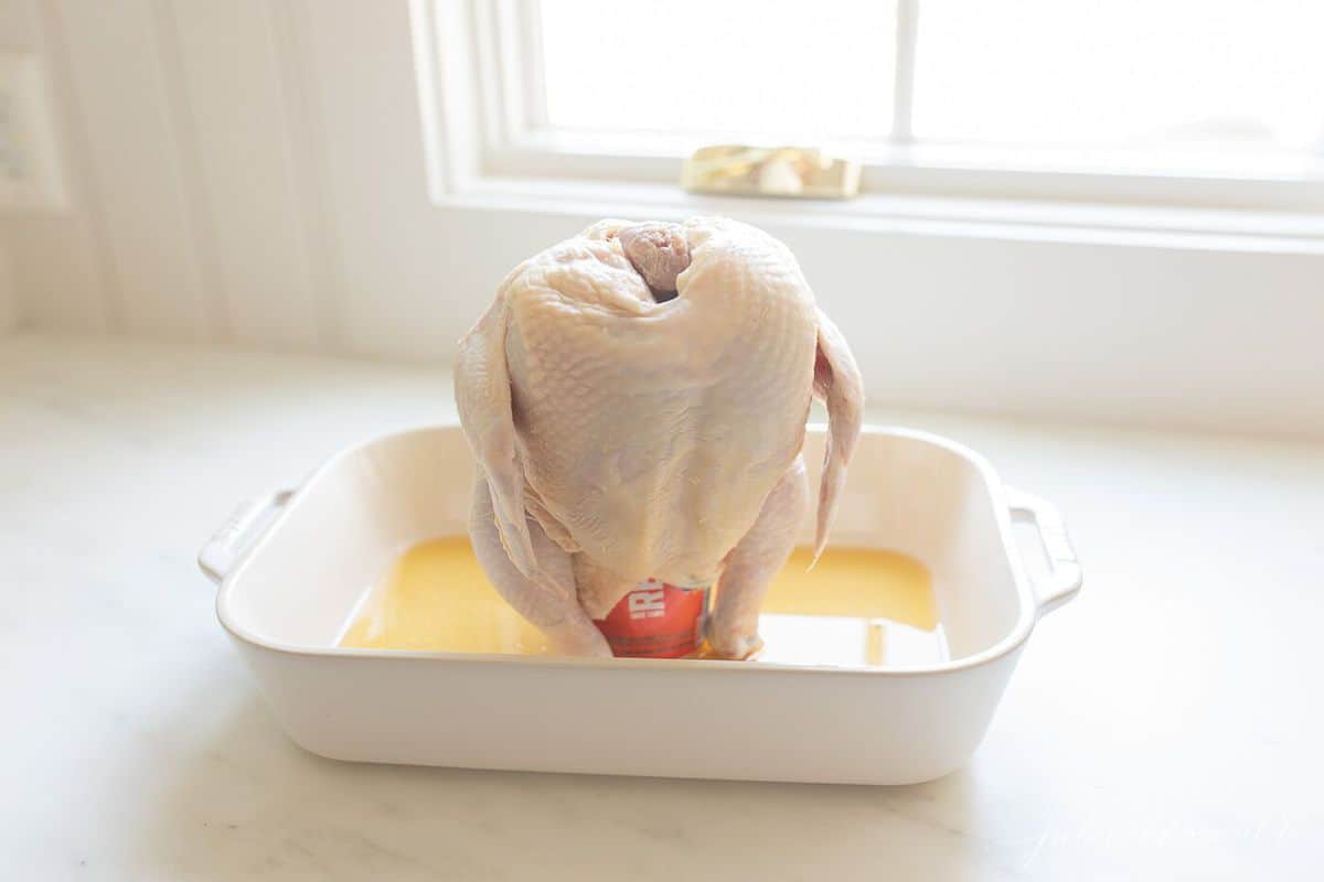 Raw whole chicken in a white roasting pan, resting on a beer can with beer at the base of the pan.