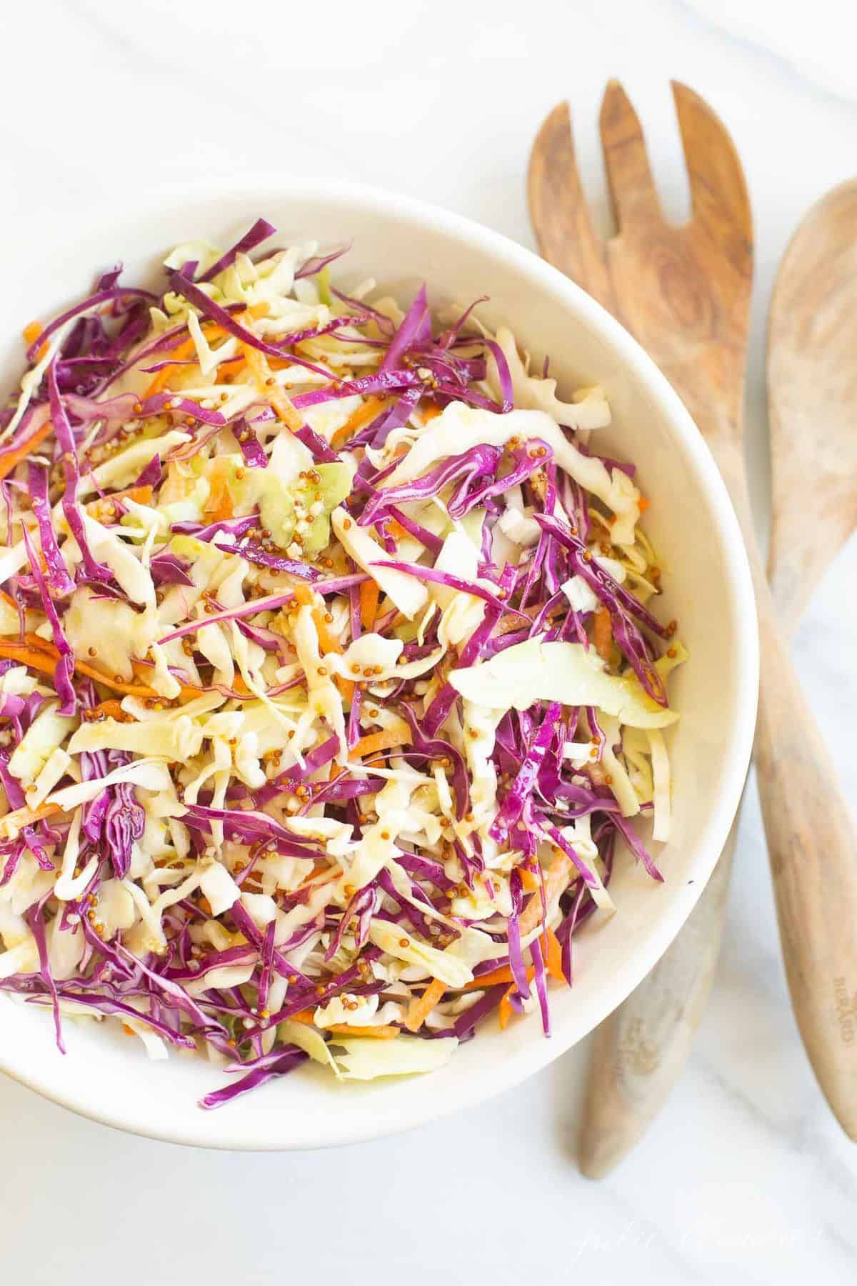 White salad bowl filled with colorful cabbage salad and wooden serving spoons. #cabbagesalad