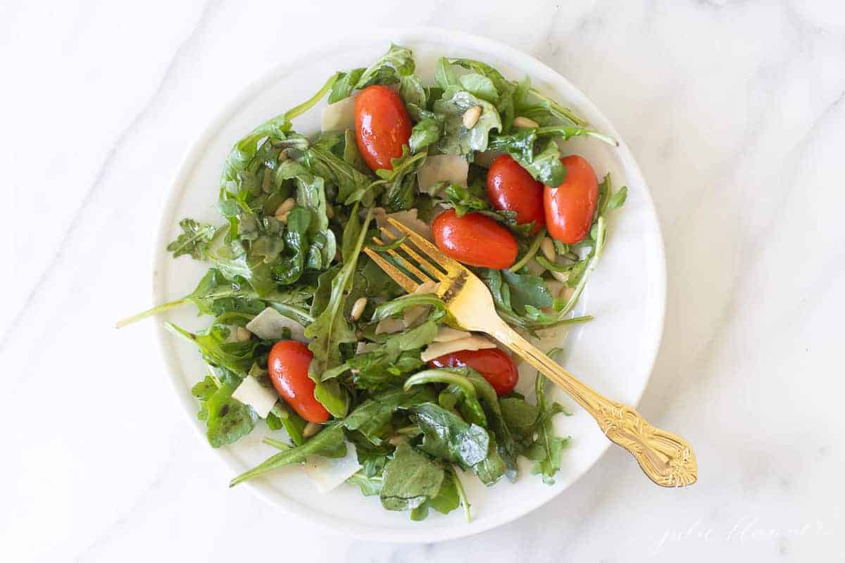 Marble countertop with a white salad plate filled with a green salad, cherry tomatoes, parmesan and more.