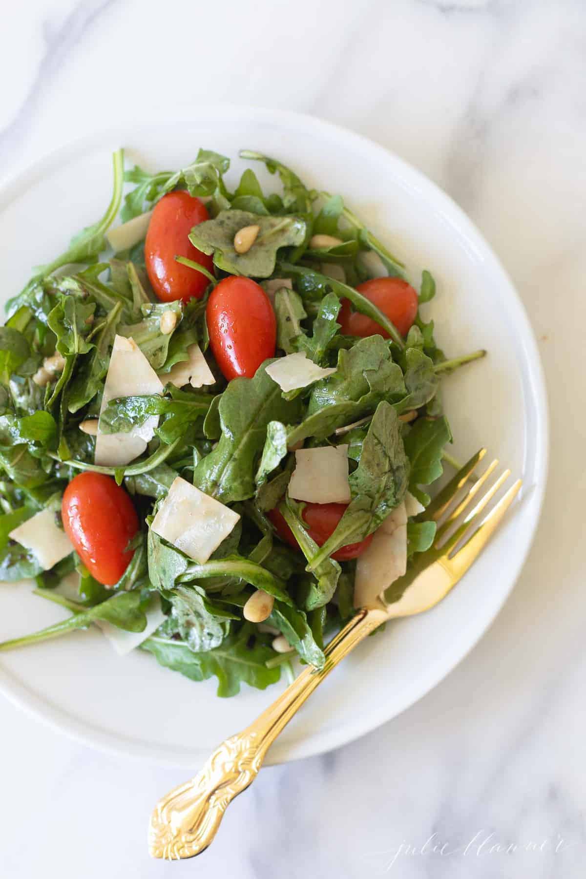 Marble countertop with a white salad plate filled with a green salad, cherry tomatoes, parmesan and more.