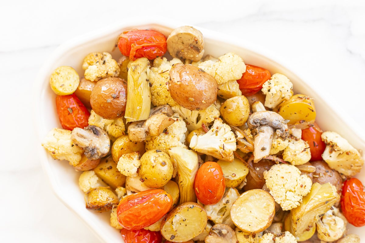Roasted Italian vegetables including potatoes, cauliflower, mushrooms, and cherry tomatoes in a white dish on a marble background.