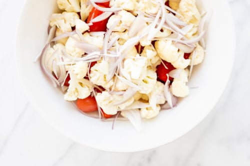 A bowl of cauliflower salad with sliced red tomatoes and onions, featuring Italian vegetables, on a white background.