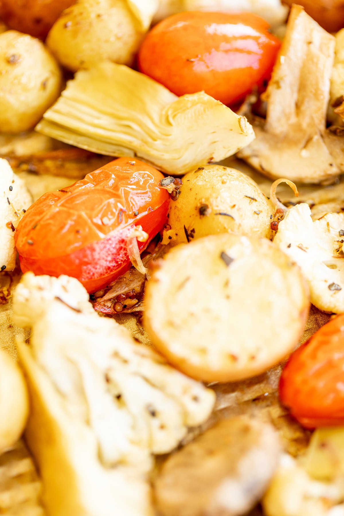 Close-up image of Italian roasted vegetables including potatoes, tomatoes, and artichokes on a baking sheet.