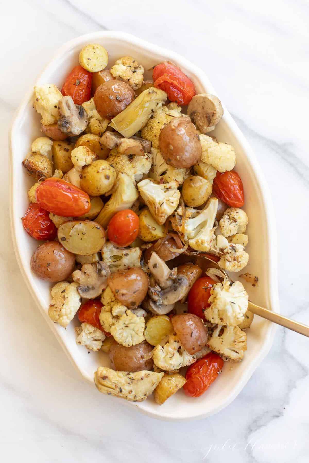 Ivory oval ceramic bowl filled with roasted Italian vegetables, gold serving spoon to the side.