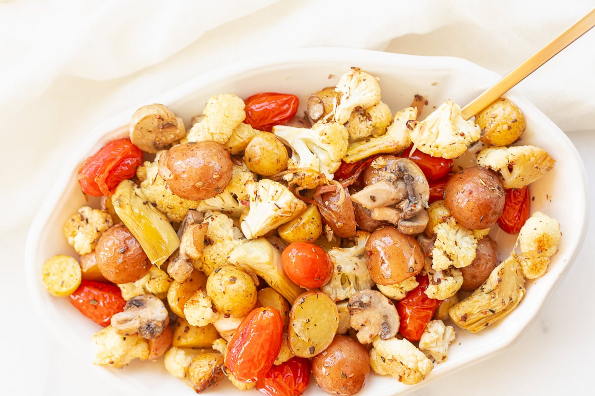 Roasted Italian vegetables including potatoes, cauliflower, mushrooms, and tomatoes in a white dish with a gold fork.