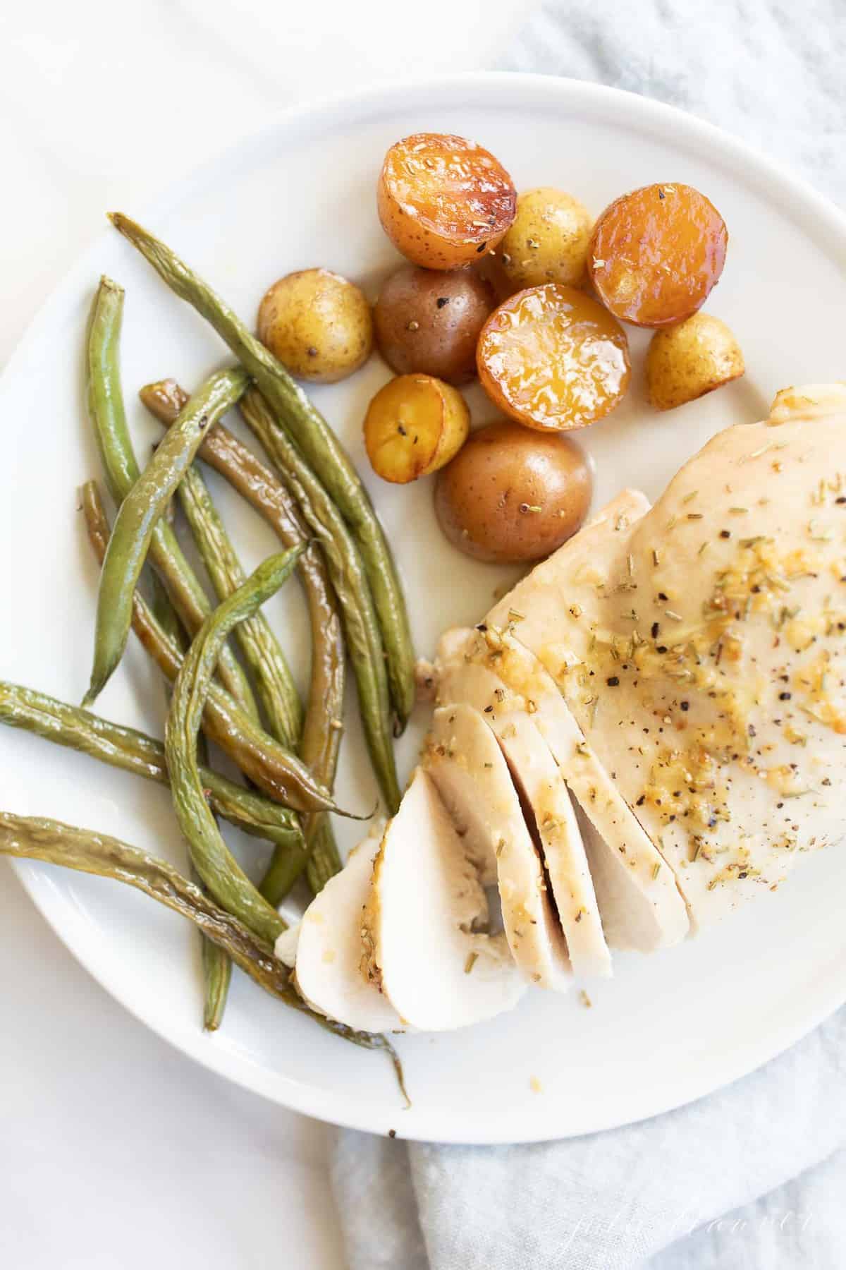 White plate close-up, filled with baked rosemary chicken, green beans and baby potatoes. #rosemarychicken #lemonrosemarychicken #bakedchickenandveggies