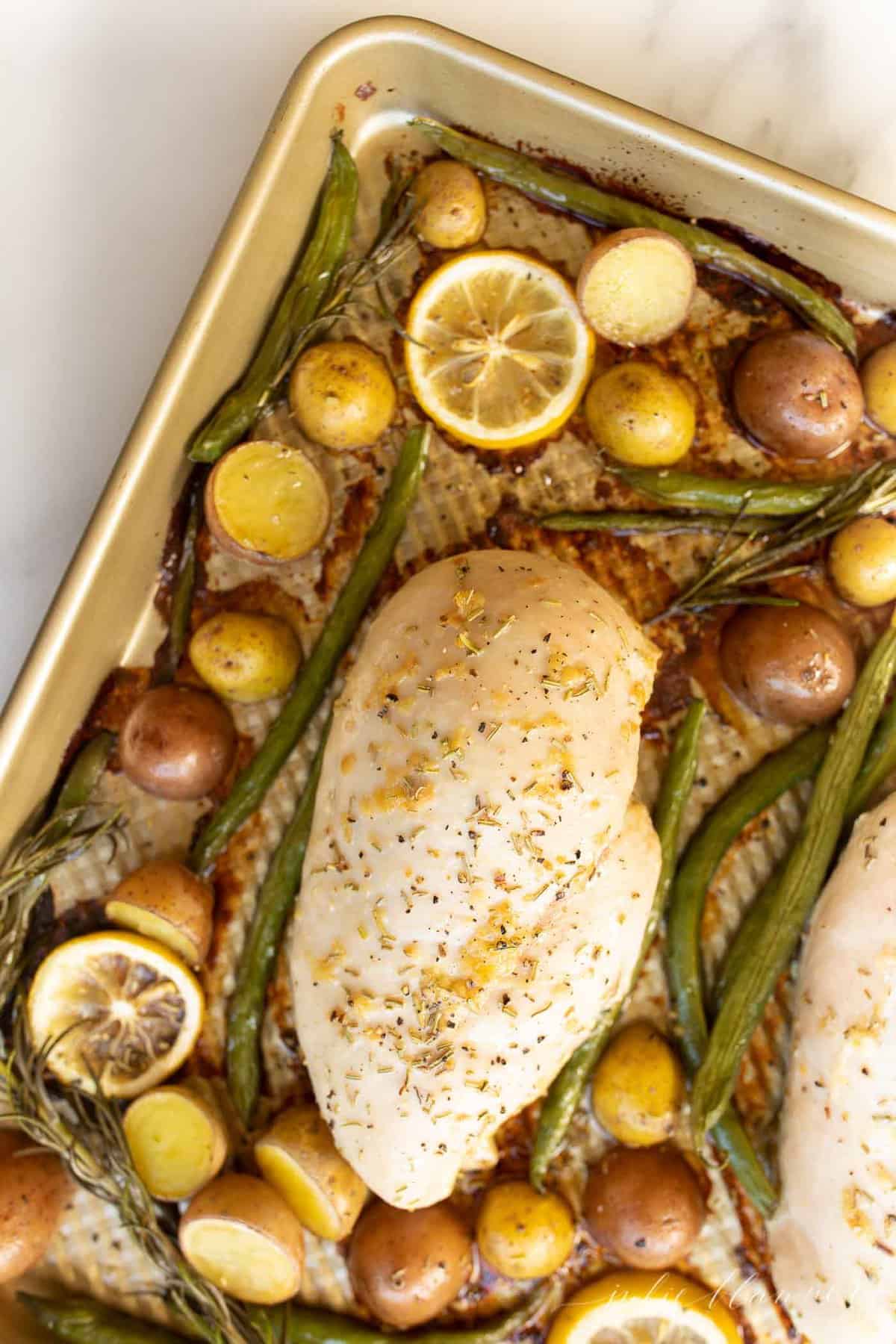 Gold sheetpan filled with baked chicken and veggies such as green beans, baby potatoes and lemon slices. #bakedchickenandveggies #rosemarychicken