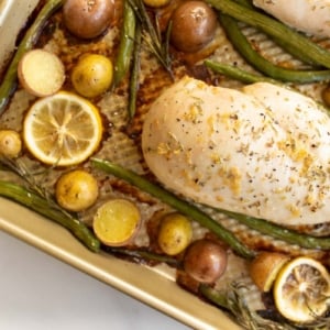 Gold sheet pan filled with baked chicken and veggies such as green beans, baby potatoes and lemon slices.