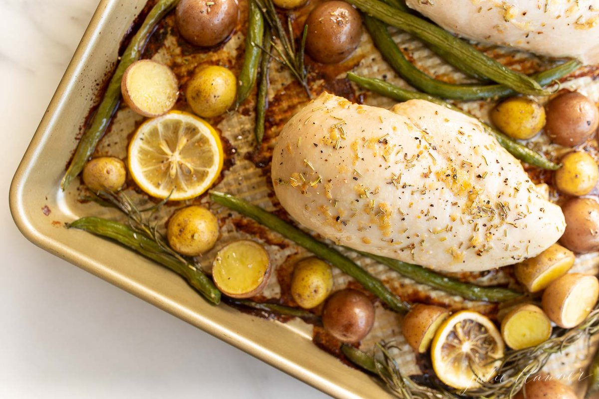 Gold sheet pan filled with baked chicken and veggies such as green beans, baby potatoes and lemon slices.