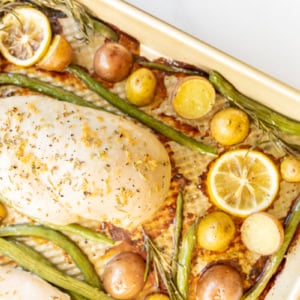 Roasted rosemary chicken breasts with lemon slices, green beans, and potatoes on a baking sheet.