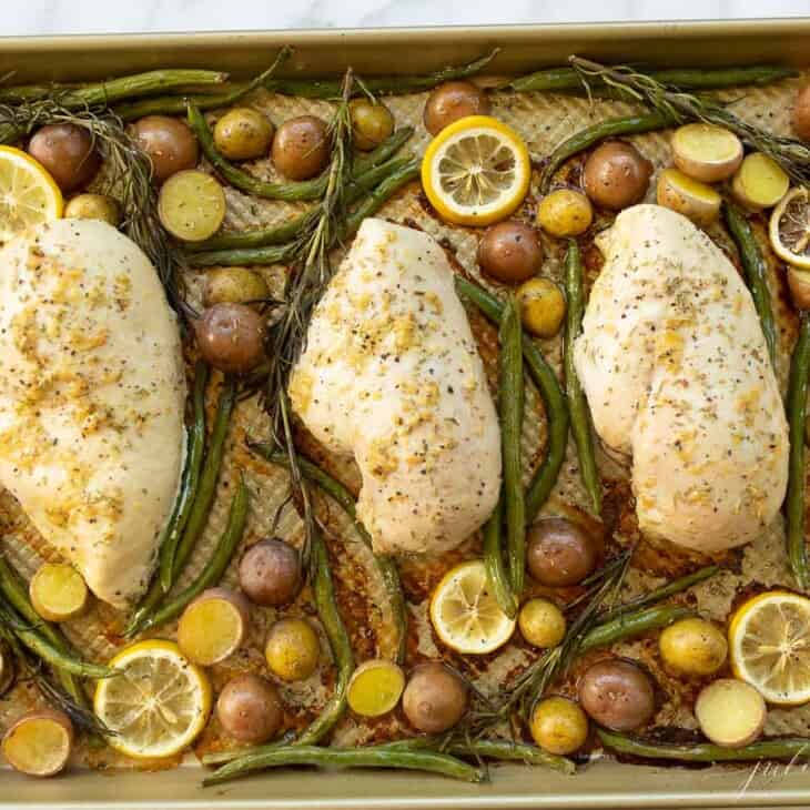 Gold sheetpan filled with baked chicken and veggies such as green beans, baby potatoes and lemon slices.