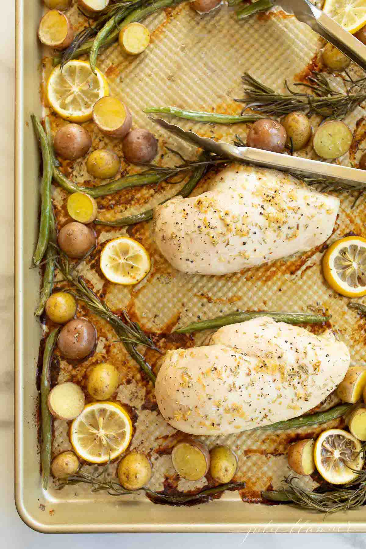 Gold sheet pan filled with baked chicken and veggies such as green beans, baby potatoes and lemon slices.