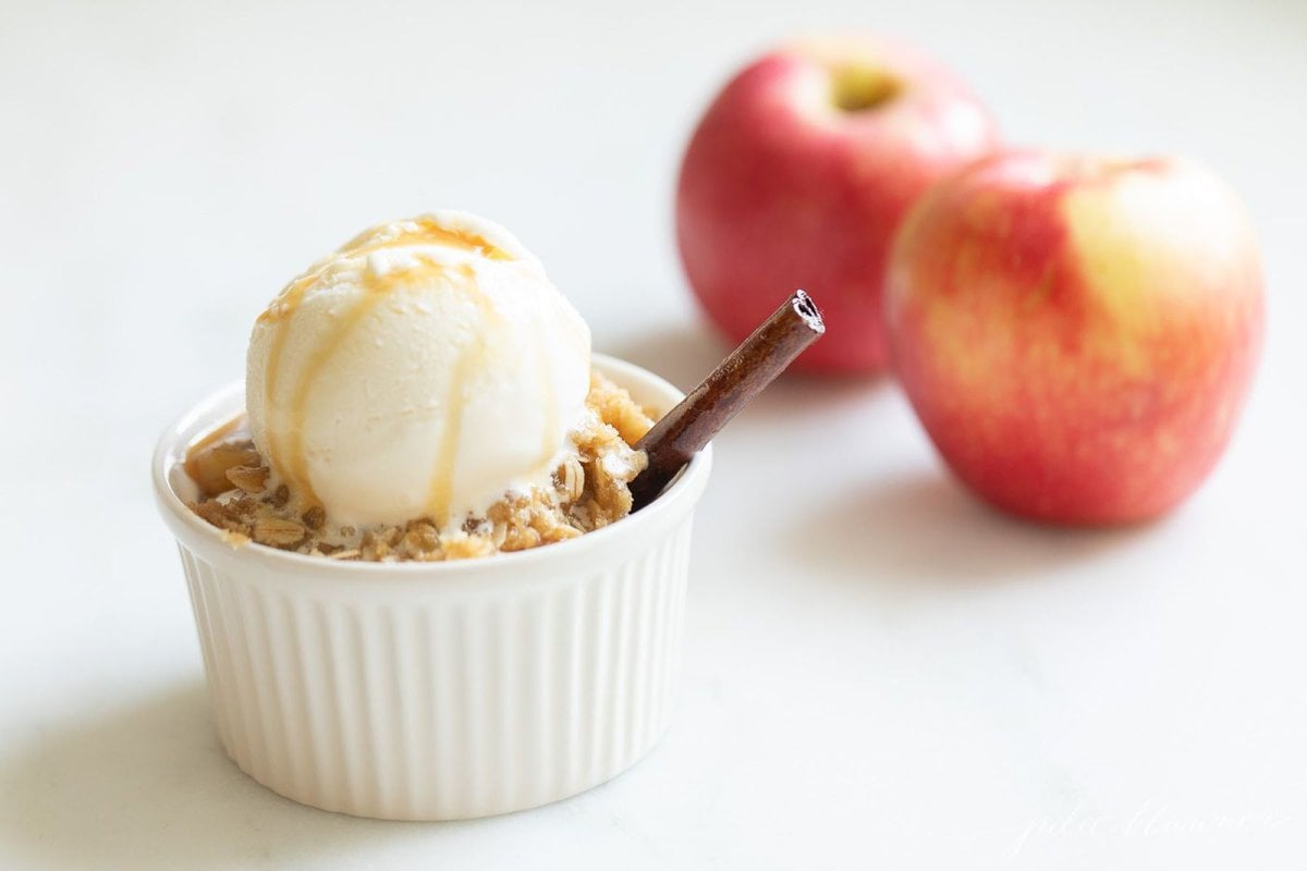 Apple crisp topped with ice cream in a small white ramekin, apples in the background