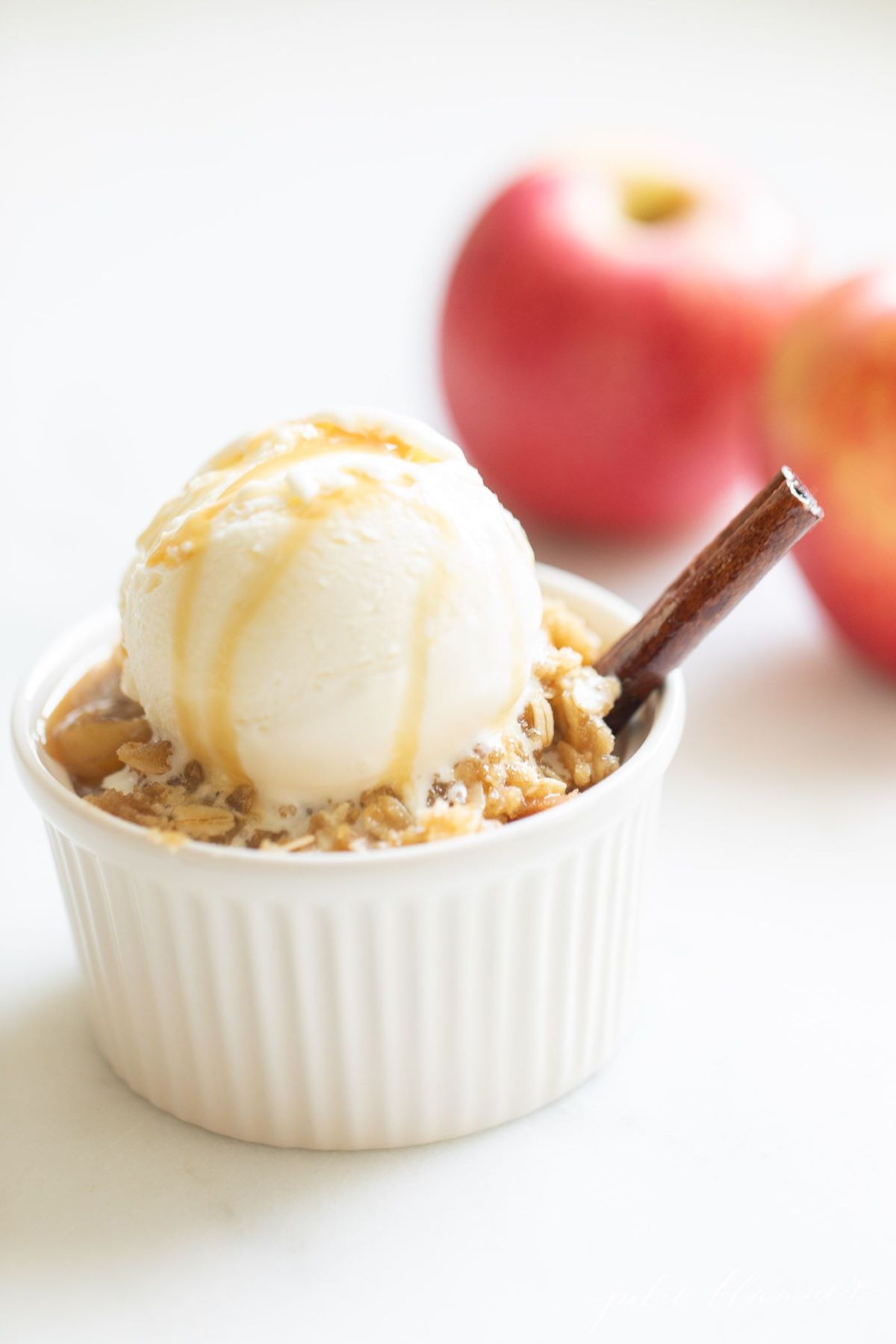 Apple crisp topped with ice cream in a small white ramekin, apples in the background