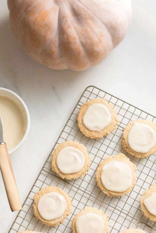 A bowl of frosting and a pumpkin to the side of sugar cookies cooling on a cooling rack.