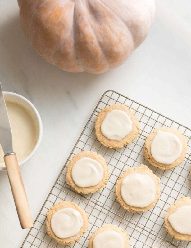 A bowl of frosting and a pumpkin to the side of sugar cookies cooling on a cooling rack.
