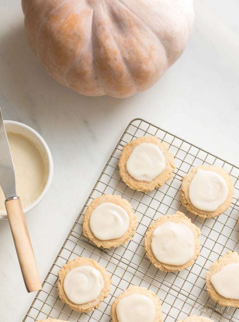 A bowl of frosting and a pumpkin to the side of sugar cookies cooling on a cooling rack. 