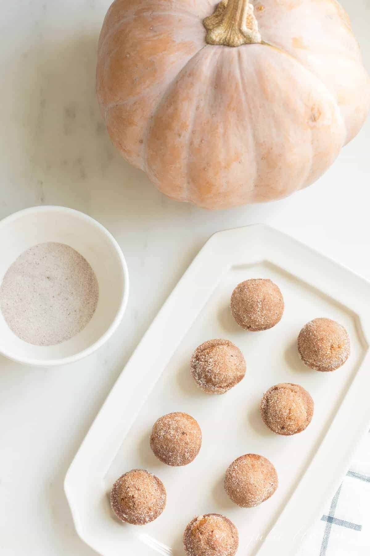 A pumpkin and bowl of cinnamon sugar to the side of a white tray with mini muffins on top.
