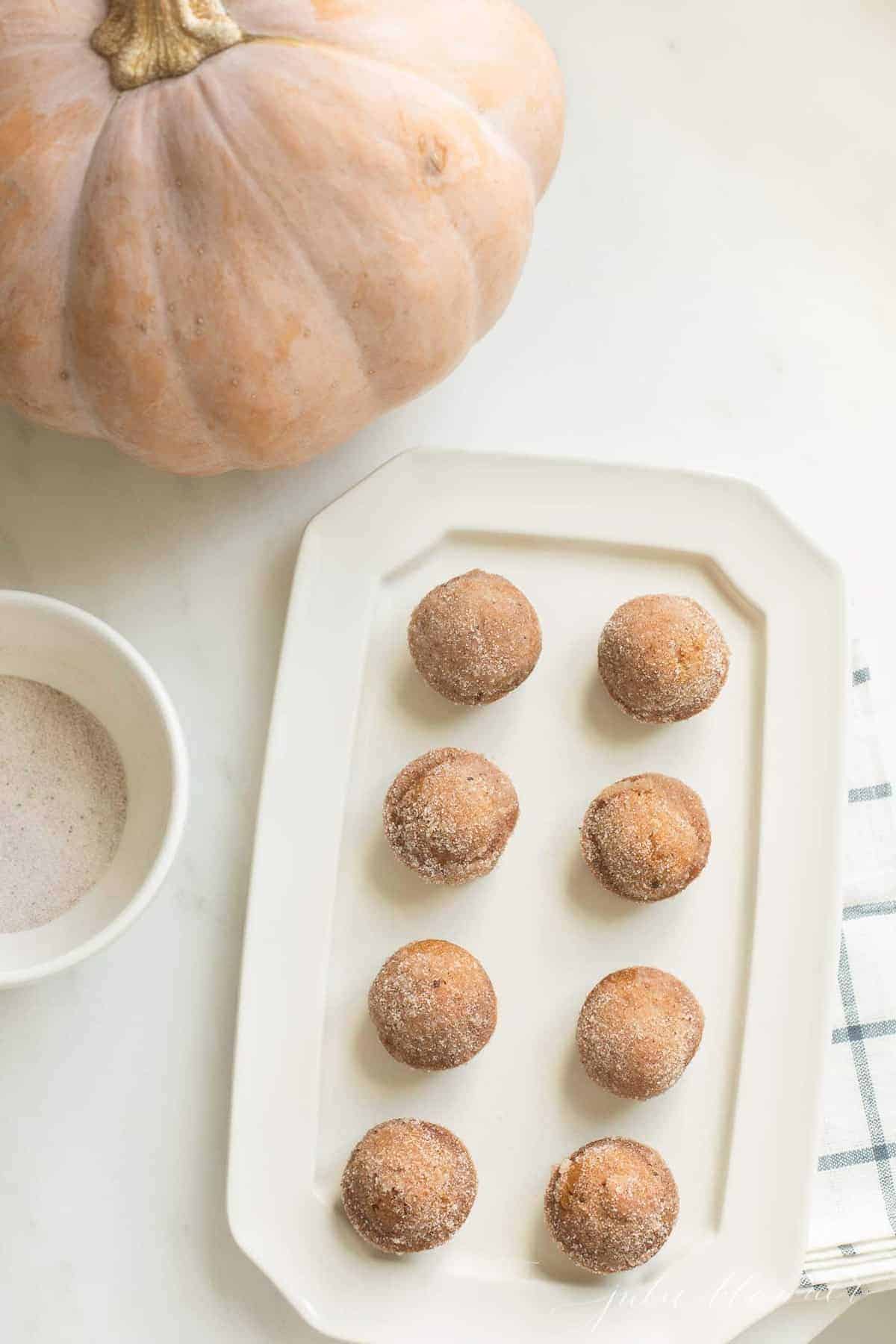 A pumpkin and bowl of cinnamon sugar to the side of mini muffins on a white platter. 