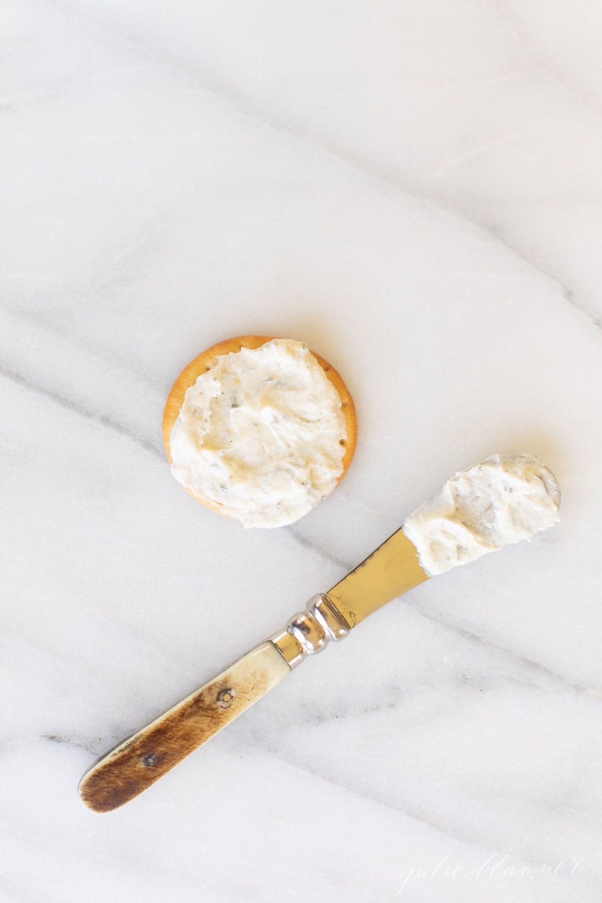 A spreader knife, spreading homemade Boursin cheese on a cracker.