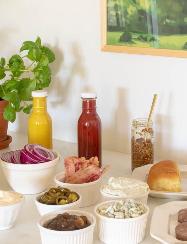 Burger toppings displayed in white ceramic bowls, condiments in rear and buns to the side, burgers in foreground. #burgerbar #burgershop #sliderbar