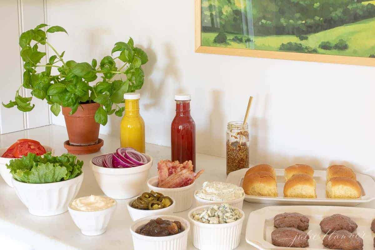 Burger toppings displayed in white ceramic bowls, condiments in rear and buns to the side, burgers in foreground. #burgerbar #burgershop #sliderbar