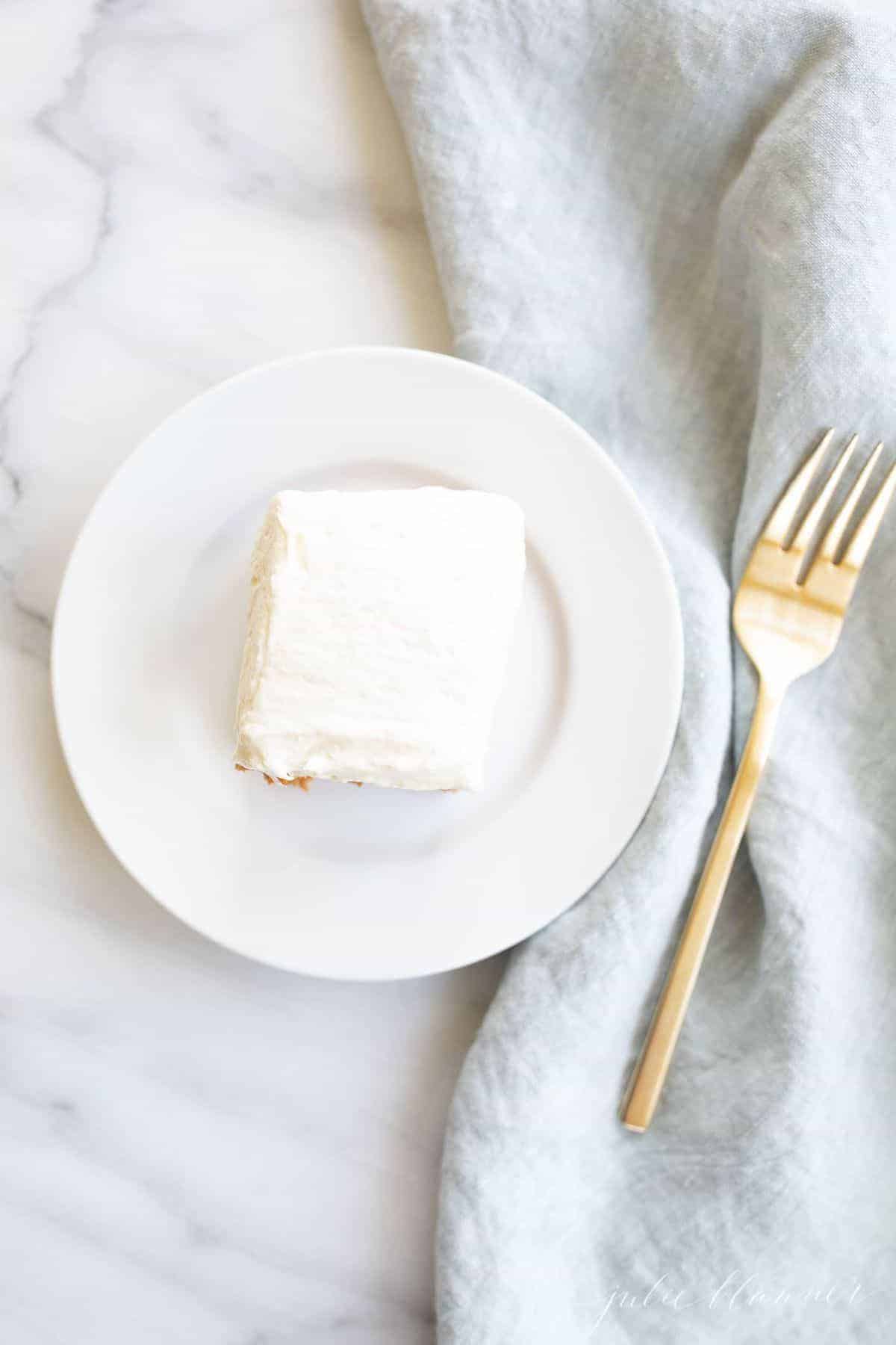 A piece of cake, frosted with fluffy cream cheese frosting on a marble background. Blue linen napkin and gold fork to the side. 