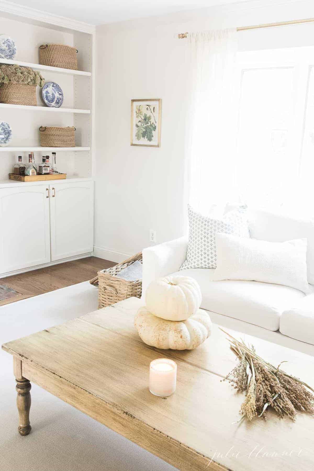 Living room featuring a white sofa, build in bookshelves, and fall decor such as a candle, fall foliage and white pumpkins on coffee table in foreground. #fallliviingroom #harvestdecor