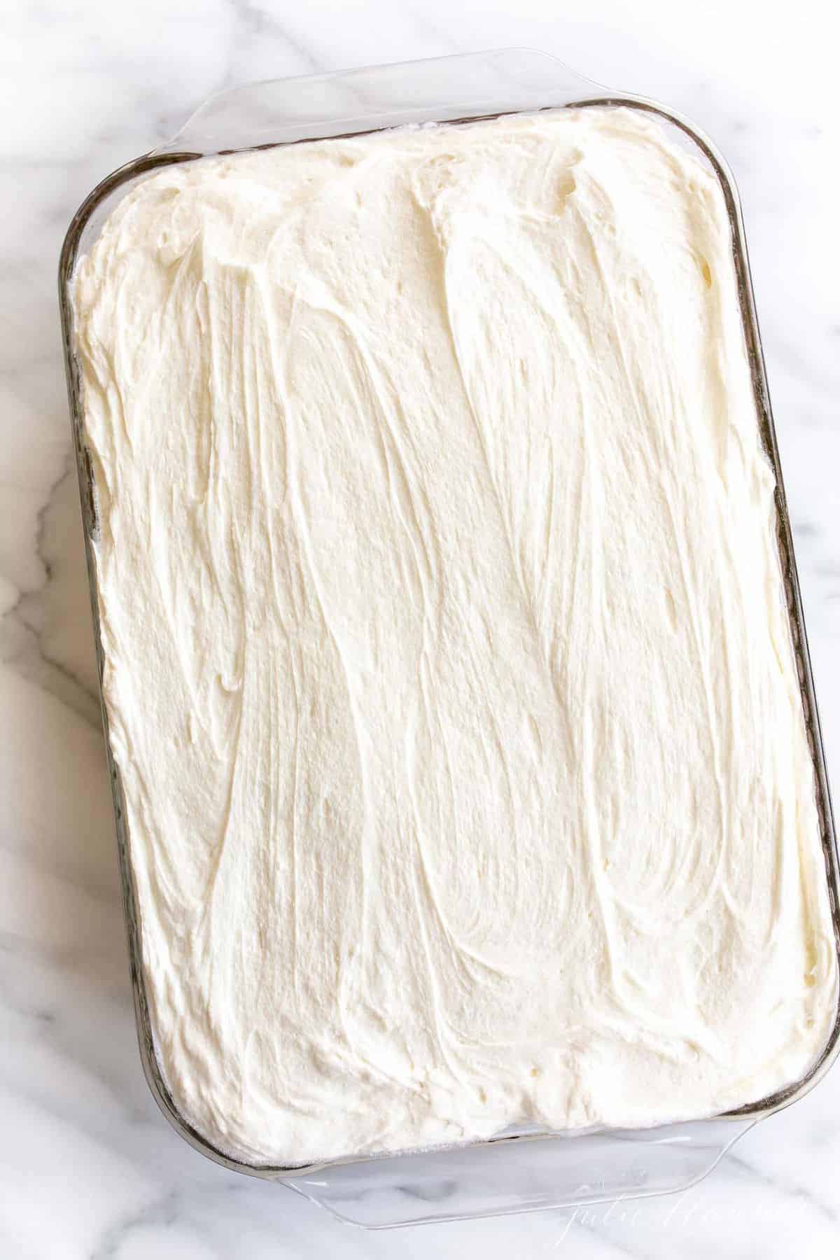 A cake frosted with cream cheese frosting in a glass dish, on a marble surface. 