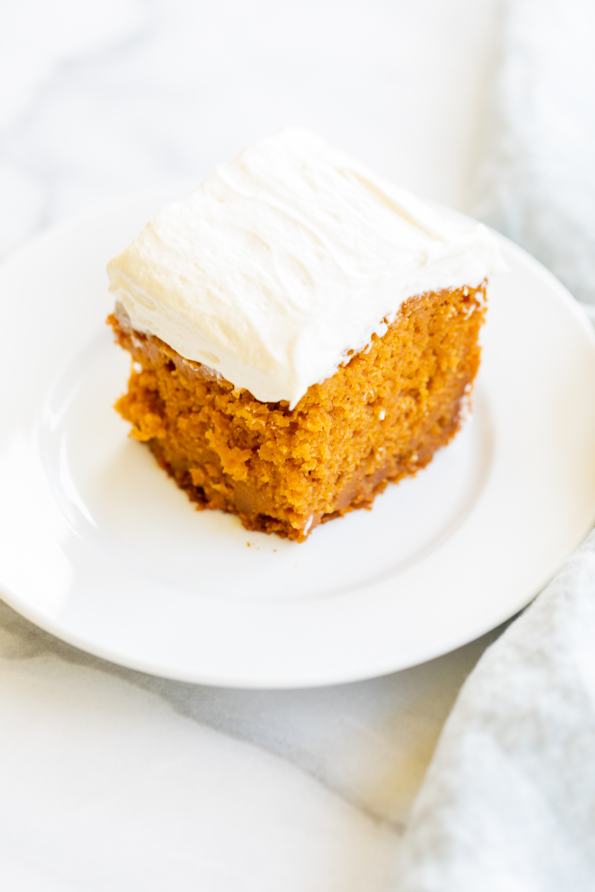 A slice of pumpkin cake with fluffy cream cheese frosting on a white plate.