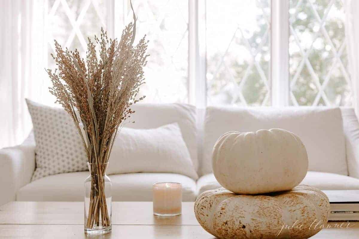 Wooden coffee table with white sofa and window in background, white pumpkins, candle and vase of fall foliage as decor. #falllivingroom #harvestdecor