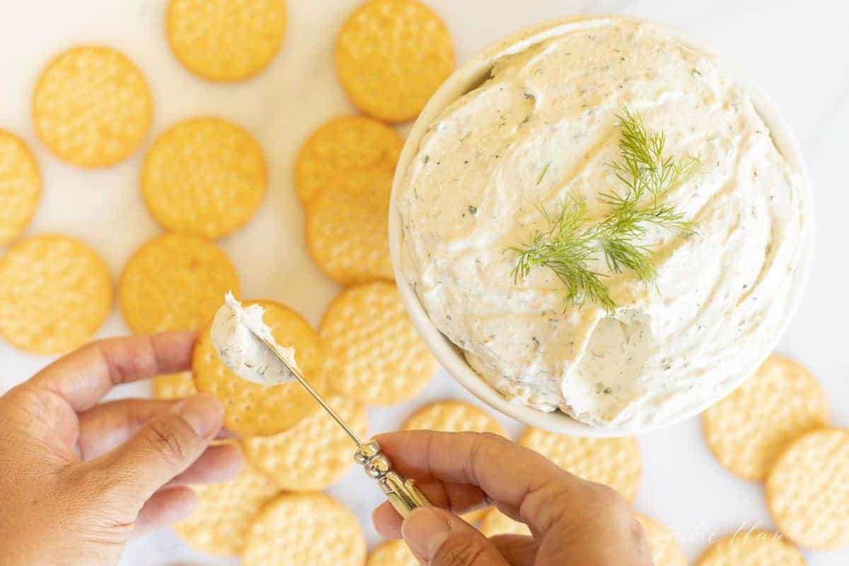 White bowl of herb cream cheese with crackers, and a hand spreading cheese onto a cracker. #homemadeboursin 