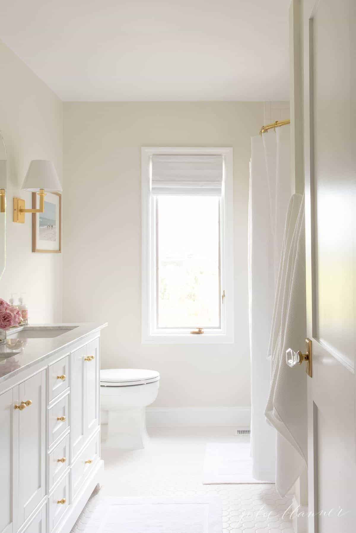 A look into a white bathroom with a door slightly ajar. Gold finishes and pink flowers on the vanity. #spabath