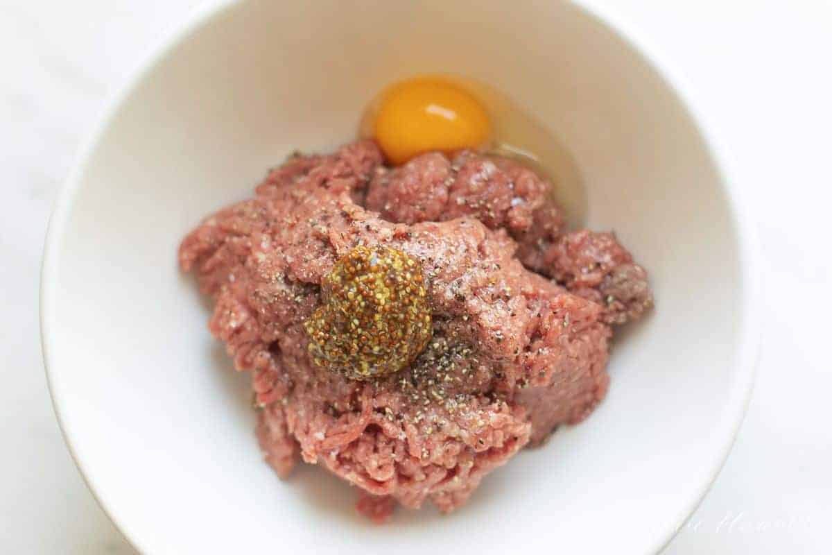 A white bowl filled with ground beef, a raw egg and seasonings. #burgersintheoven