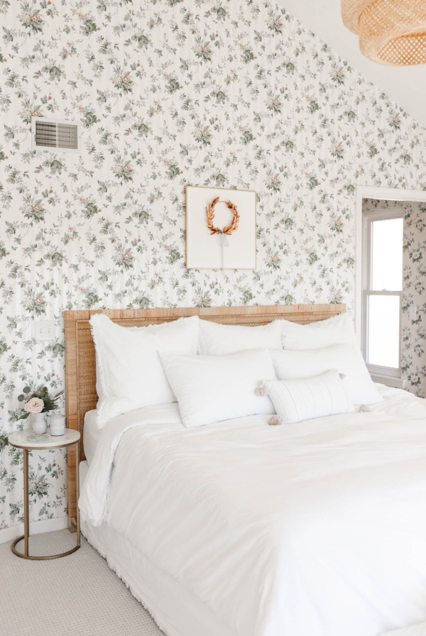 A white bed in a bedroom with floral wallpaper and a gold laurel wreath hanging above.