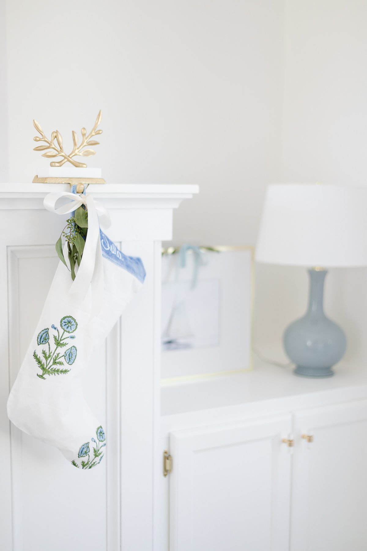 A white christmas stocking hanging on a white dresser adorned with a laurel wreath.