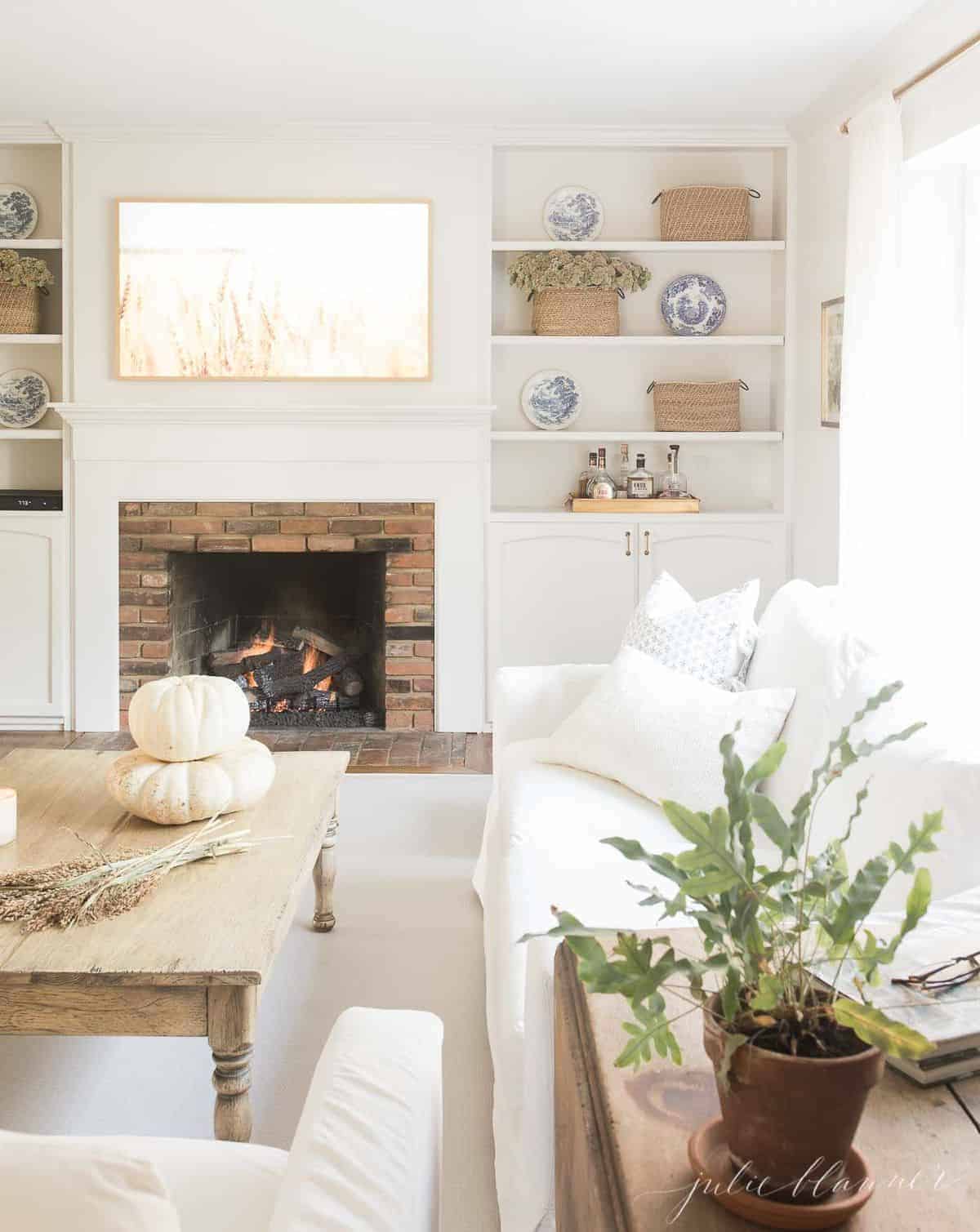 Pulled back view into a living room decorated for fall, pumpkins and wheat on coffee table and fireplace burning in background. #fallharvestdecor