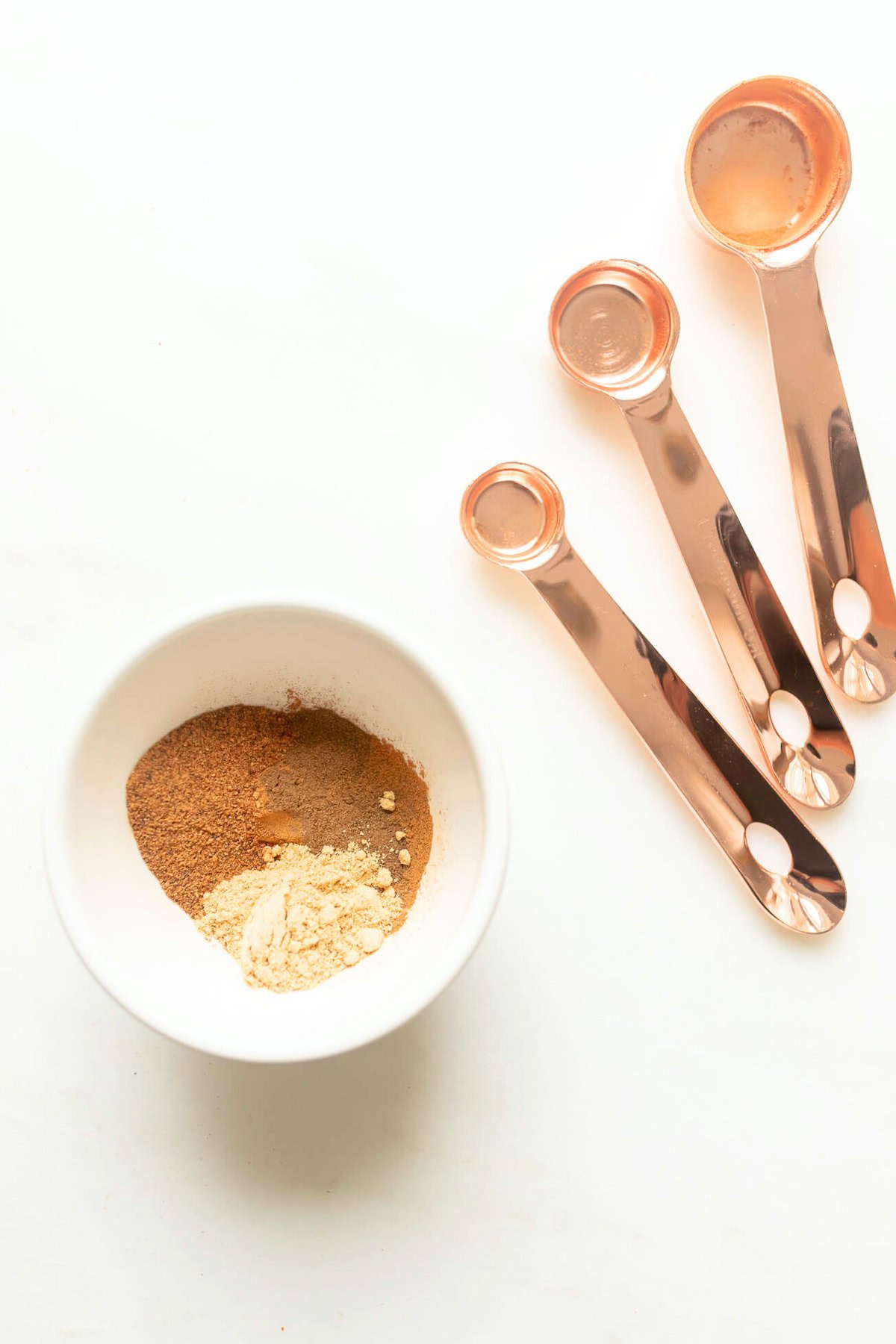 Three measuring spoons of varying sizes filled with ground cinnamon on a white background, next to a bowl of spices.