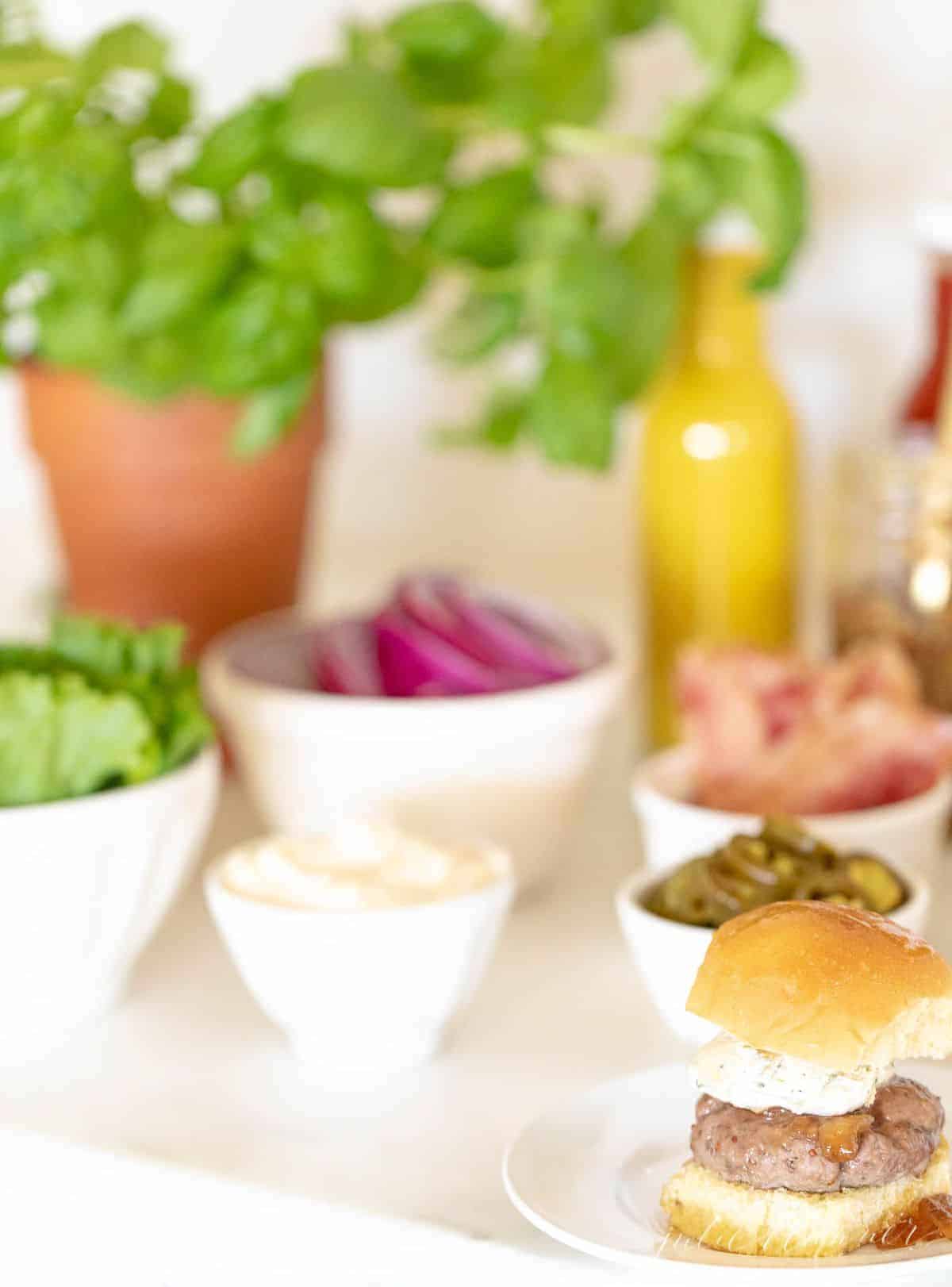 Burger toppings displayed in white ceramic bowls, slider in foreground. #burgerbar #burgershop #sliderbar