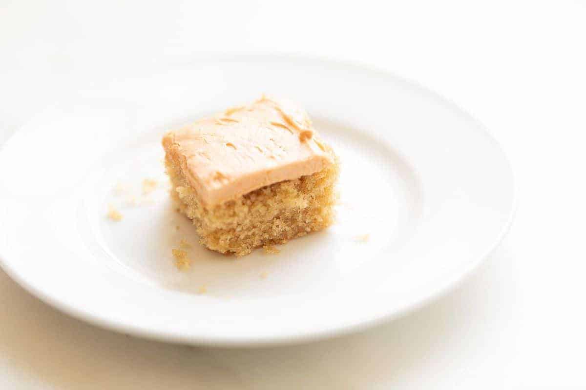 A piece of frosted Butterscotch cake on a white plate, crumbs surrounding it. 