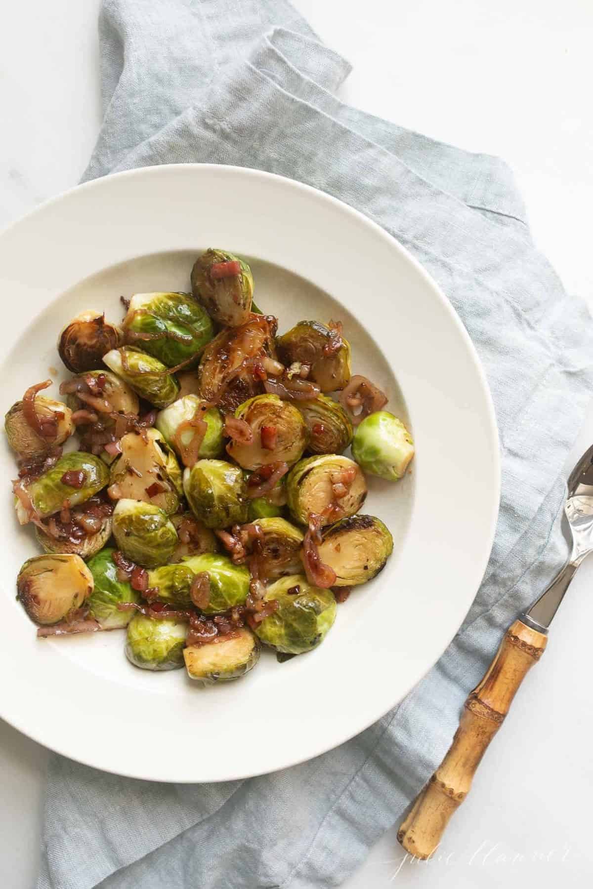Crispy brussel sprouts on a white plate with a blue linen napkin and serving spoon to the side. 