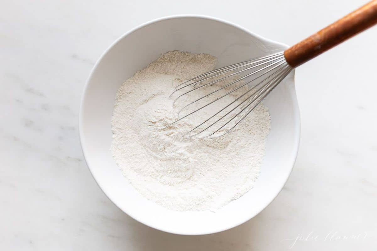 Flour with a whisk inside a white bowl on a white surface.